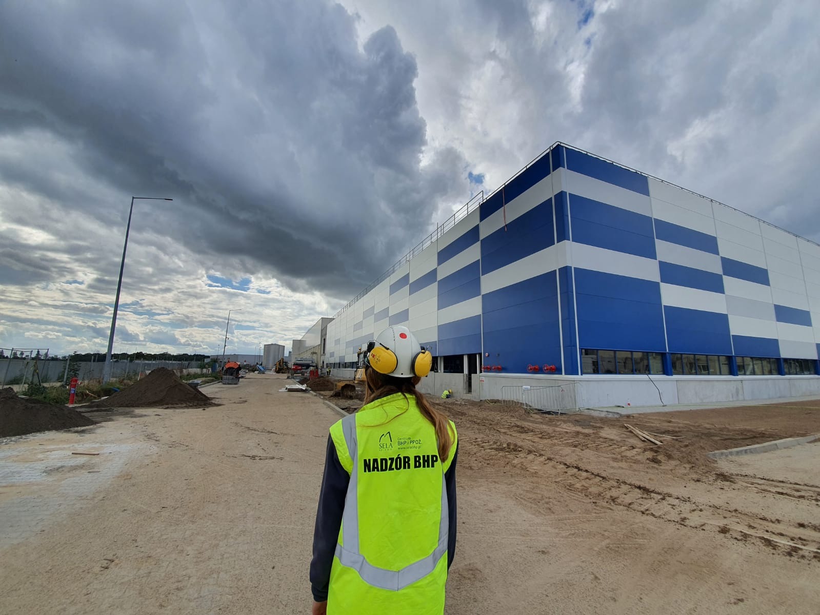 Osoba w kamizelce odblaskowej z napisem 'Nadzór BHP' i kasku ochronnym na tle budowy hali magazynowej z niebiesko-białą elewacją w kratkę.