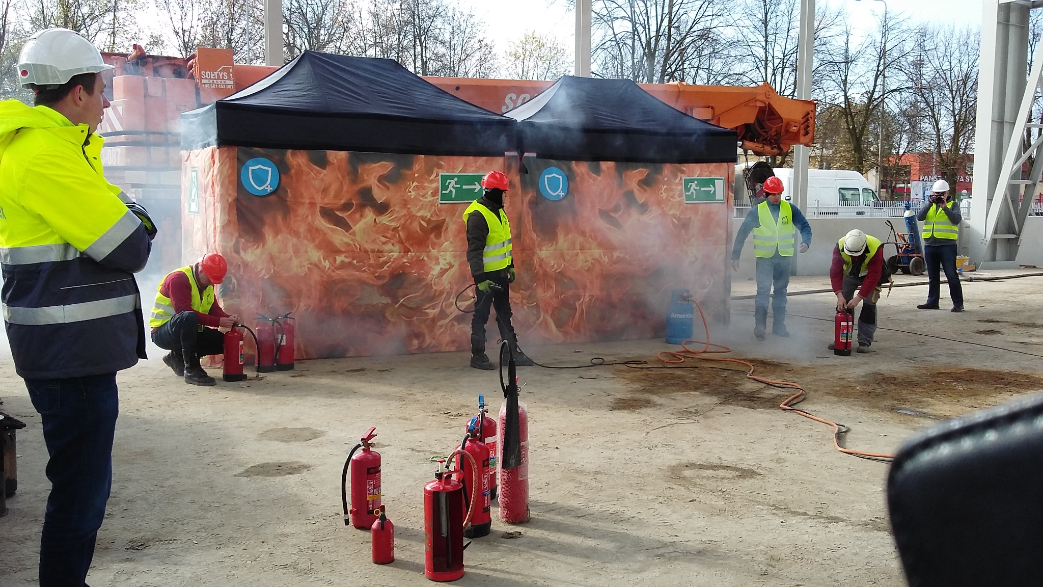 Grupa osób w kaskach i kamizelkach odblaskowych ćwiczy gaszenie pożaru przy użyciu gaśnic, w tle namioty z grafiką płomieni i strzałką ewakuacyjną.