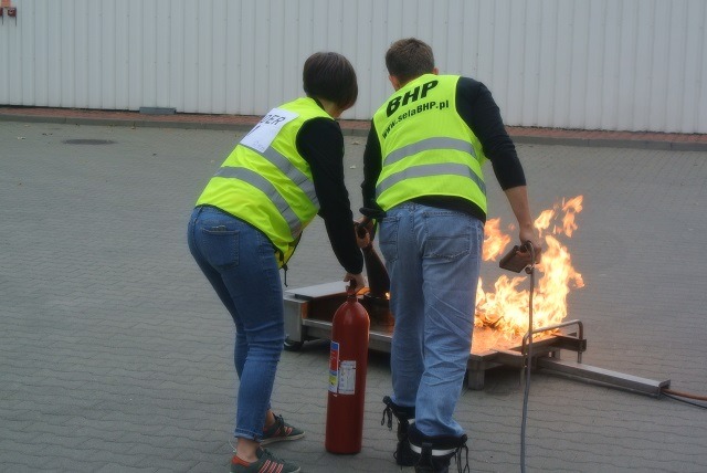 Dwie osoby w odblaskowych kamizelkach demonstrują gaszenie płonącego ognia przy użyciu gaśnicy podczas szkolenia.