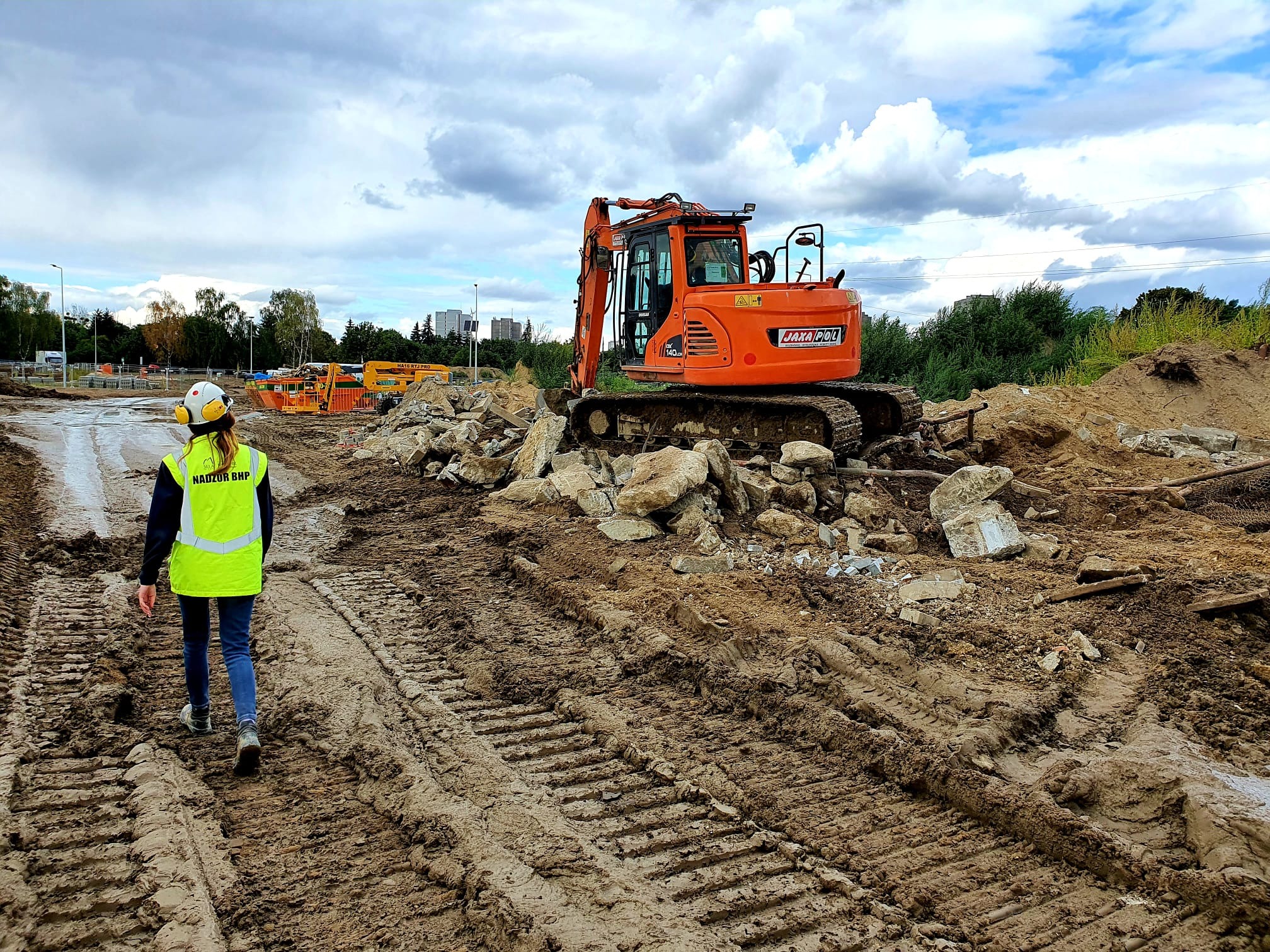 Kobieta w kamizelce odblaskowej z napisem 'Nadzór BHP' idzie po błotnistym terenie budowy w kierunku pomarańczowej koparki stojącej na gruzie.