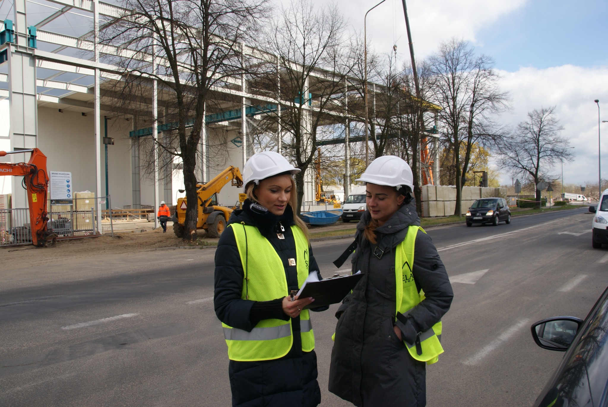 Dwie kobiety w kaskach i kamizelkach odblaskowych analizują dokumenty na tle placu budowy z ciężkim sprzętem i konstrukcją stalową.