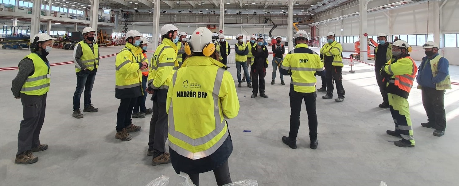 Grupa osób w odblaskowych kamizelkach BHP i kaskach ochronnych na terenie hali produkcyjnej podczas instruktażu, niektóre osoby w maseczkach ochronnych.