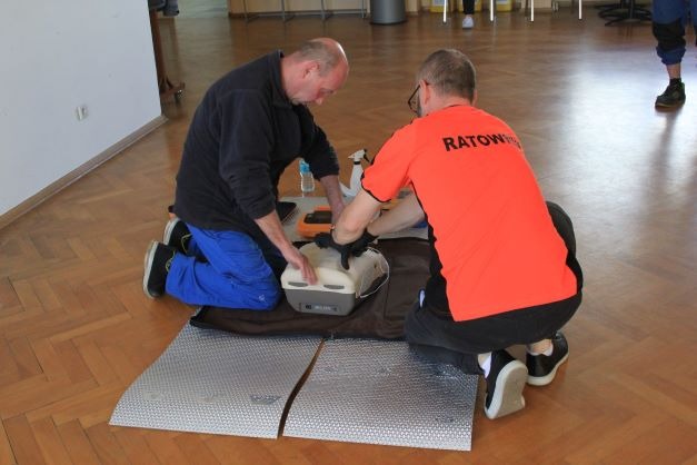 Instruktorzy demonstrują użycie defibrylatora AED na manekinie podczas zajęć, jeden w pomarańczowej koszulce z napisem 'RATOWNIK'.
