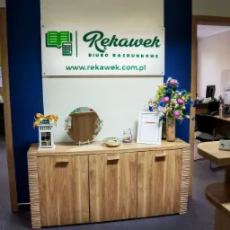 Recepcja biura z logo firmy, kwiatami i elementami dekoracyjnymi, widok na wnętrze biura.