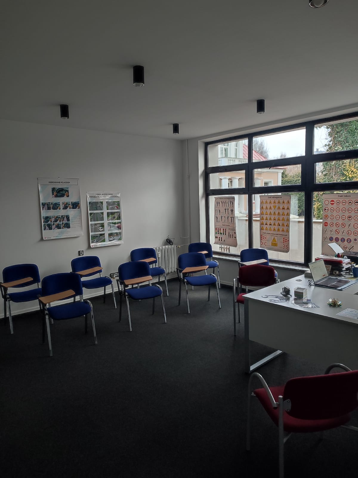 Sala wykładowa z niebieskimi krzesłami, biurkiem z laptopem i plakatami ze znakami drogowymi na ścianie przy oknie.