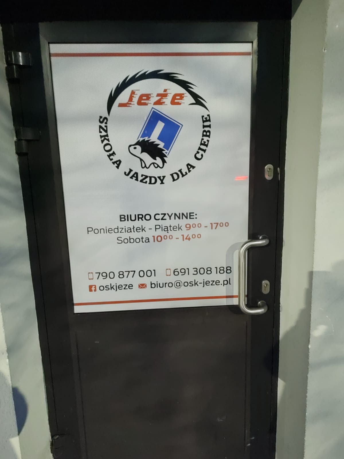 Wejście do biura z logo z jeżem i znakiem drogowym, informacja o godzinach otwarcia i numerach kontaktowych.