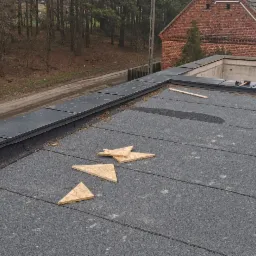 Płaski dach pokryty papą, z fragmentami obróbek blacharskich i drewnianymi klinami, widok na okoliczne budynki i drzewa.