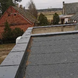 Widok z góry na płaski dach pokryty papą z obróbkami blacharskimi, w tle budynki gospodarcze i pola uprawne w pochmurny dzień.