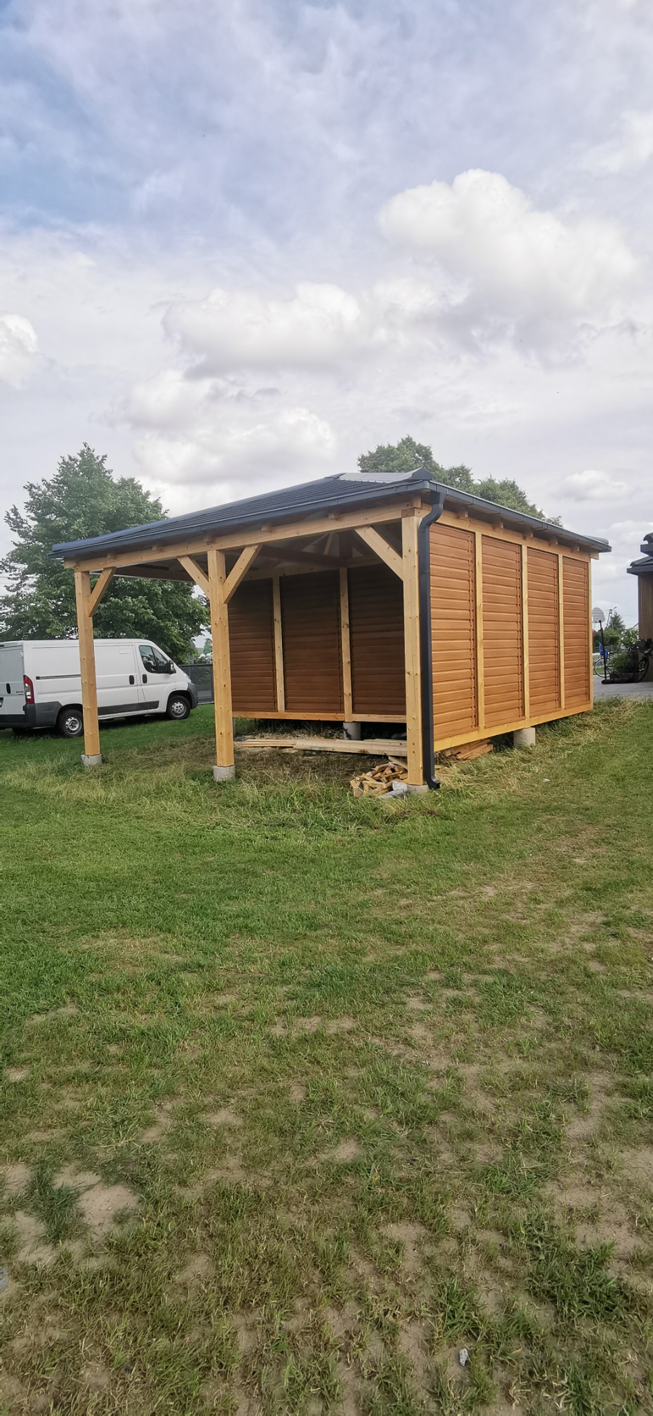 Nowa, drewniana altana z czarnym dachem i rynną, widoczna na trawniku obok białego samochodu dostawczego.