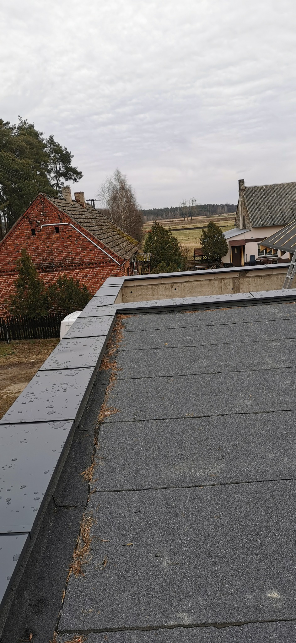 Widok z góry na płaski dach pokryty papą z obróbkami blacharskimi, w tle budynki gospodarcze i pola uprawne w pochmurny dzień.