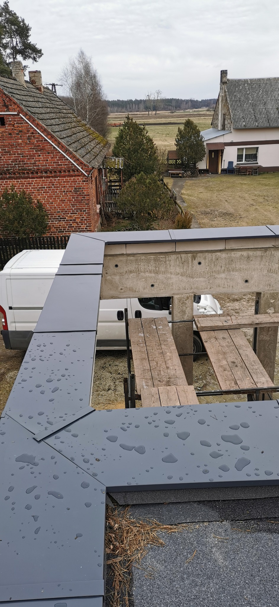 Szare, metalowe elementy wykończeniowe dachu z widocznymi kroplami wody, rusztowanie i budynki w tle.