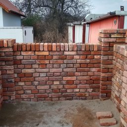 Dziedzic Maciej - Wznoszenie niskiej, murowanej konstrukcji z cegły klinkierowej, prawdopodobnie fragmentu ogrodzenia lub murku, z widocznym piaskowym podłożem i butelką wody w tle.