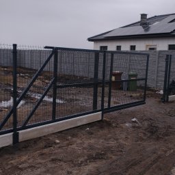 Dziedzic Maciej - Przesuwna brama wjazdowa z panelowego ogrodzenia, widoczna na tle domu z panelami słonecznymi, teren po budowie, częściowo w błocie, na pierwszym planie betonowa podstawa bramy.