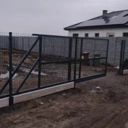 Przesuwna brama wjazdowa z panelowego ogrodzenia, widoczna na tle domu z panelami słonecznymi, teren po budowie, częściowo w błocie, na pierwszym planie betonowa podstawa bramy.