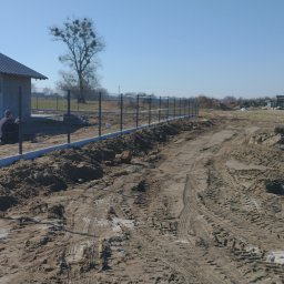 Dziedzic Maciej - Nowo zainstalowane ogrodzenie panelowe z metalową siatką na betonowej podmurówce w trakcie montażu, widoczny pracownik przy budynku, teren budowy z ziemią i śladami opon, dom w tle.