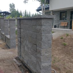 Dziedzic Maciej - Wznoszone ogrodzenie z szarej kostki betonowej, widoczny fragment muru i słupki, w tle pracownik zamiatający teren, a po prawej stronie pies obserwujący budowę.