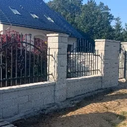 Nowoczesne ogrodzenie z metalowymi przęsłami i słupkami z szarej kostki, widok z zewnątrz posesji, dom w tle.