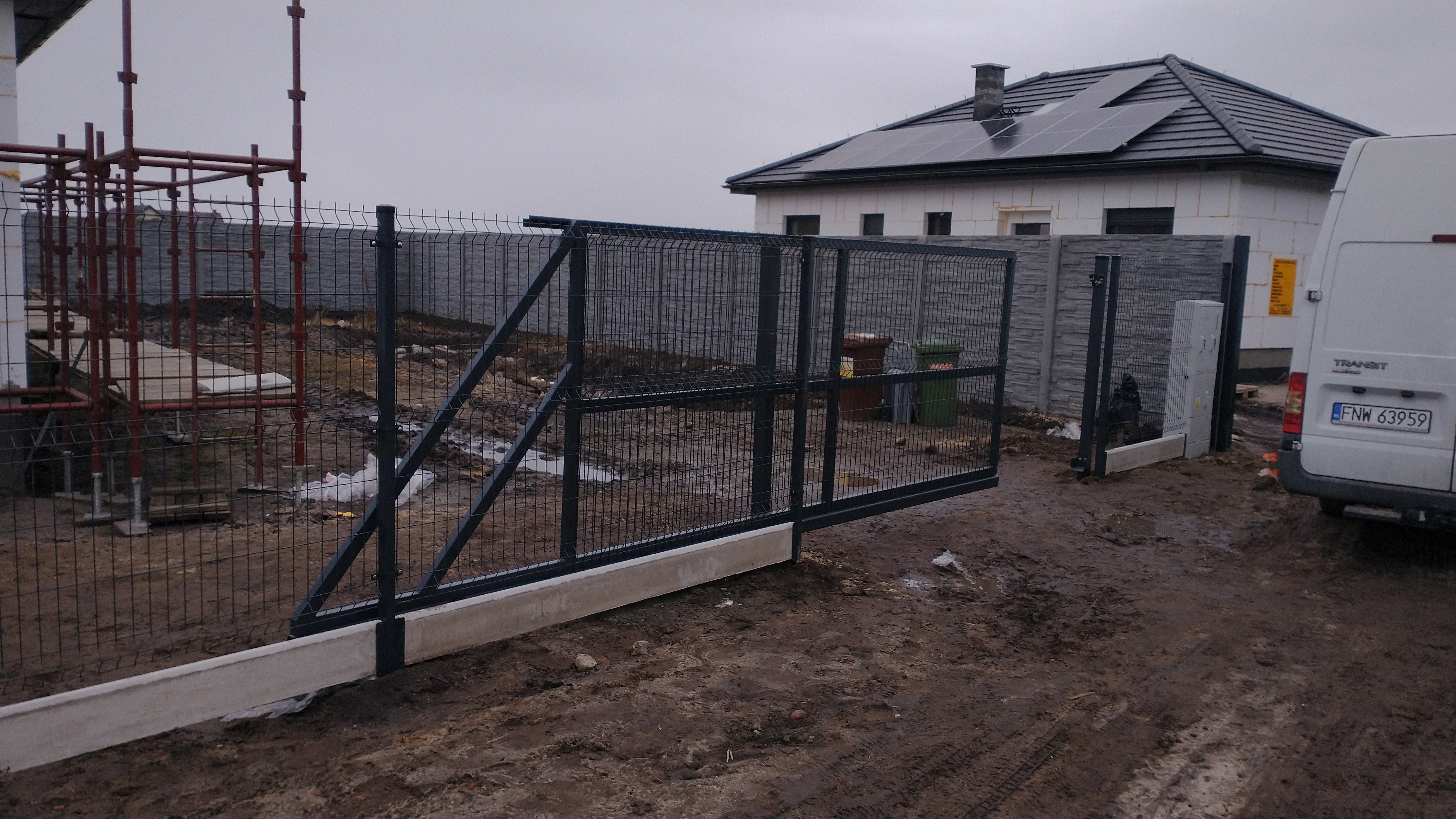 Przesuwna brama wjazdowa z panelowego ogrodzenia, widoczna na tle domu z panelami słonecznymi, teren po budowie, częściowo w błocie, na pierwszym planie betonowa podstawa bramy.