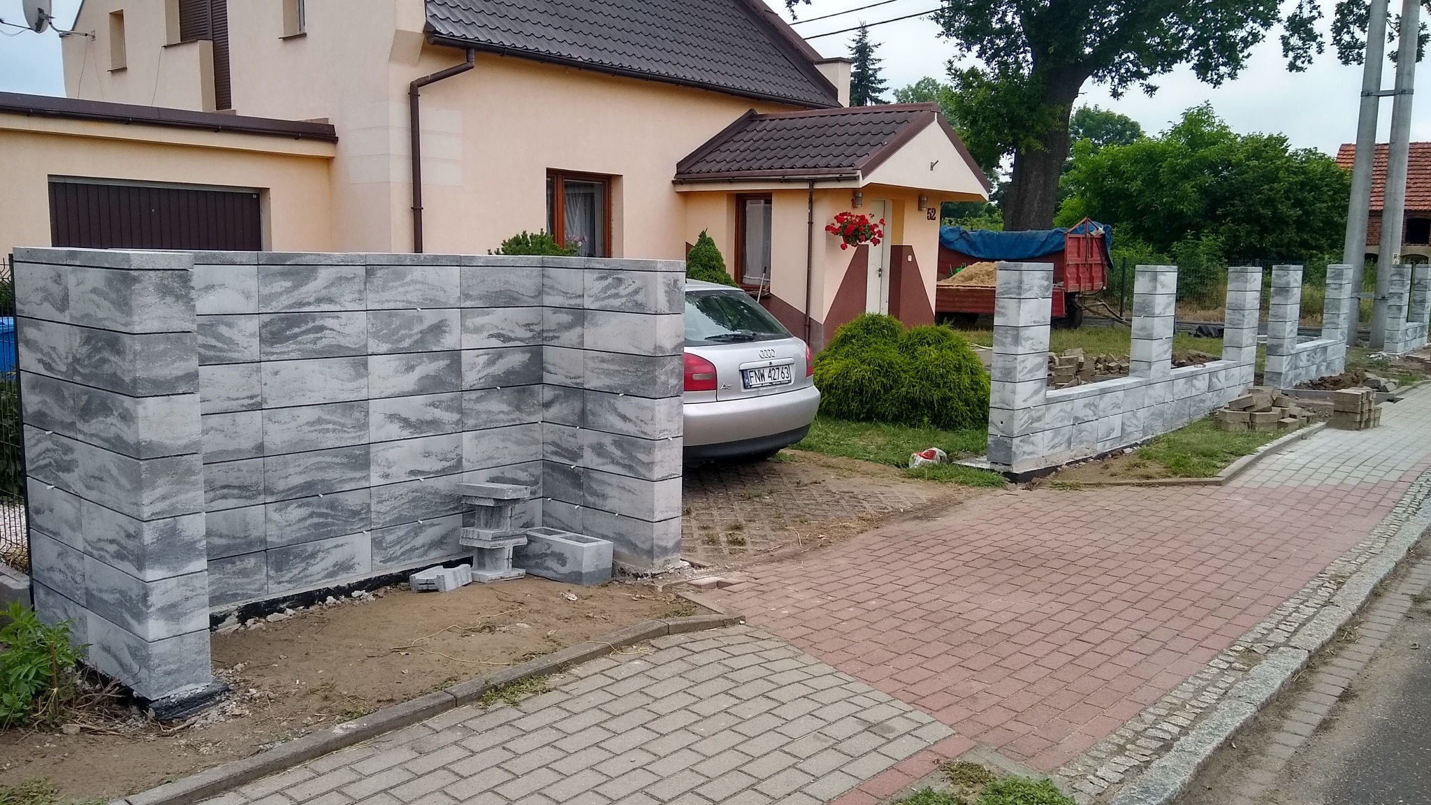 Wznoszenie ogrodzenia z szarych, wzorzystych betonowych bloczków w trakcie budowy; widoczny fragment domu z czerwonymi kwiatami w oknie, srebrny samochód i rozkopany teren.