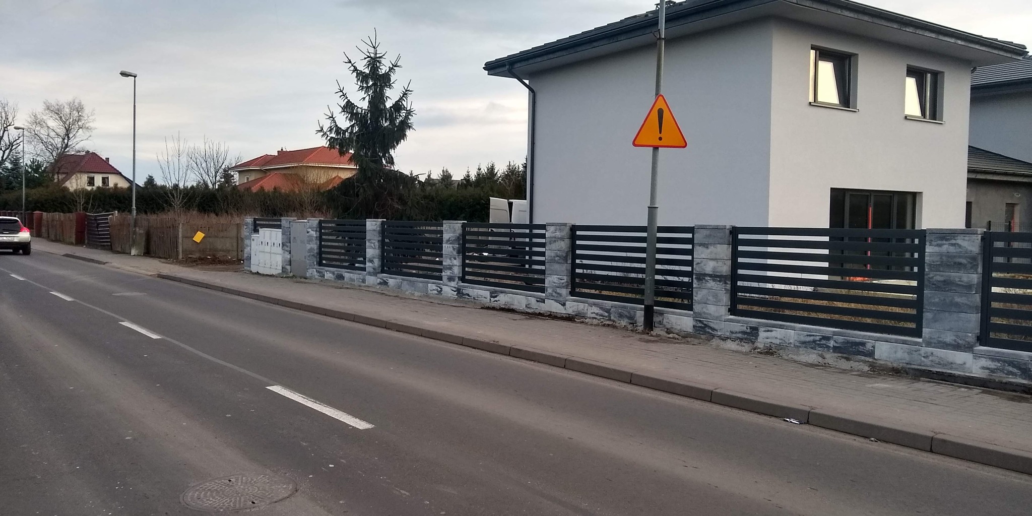 Nowoczesne ogrodzenie z poziomych, czarnych paneli metalowych i szarych, kamiennych słupków, widoczne z perspektywy ulicy z znakiem ostrzegawczym w tle.