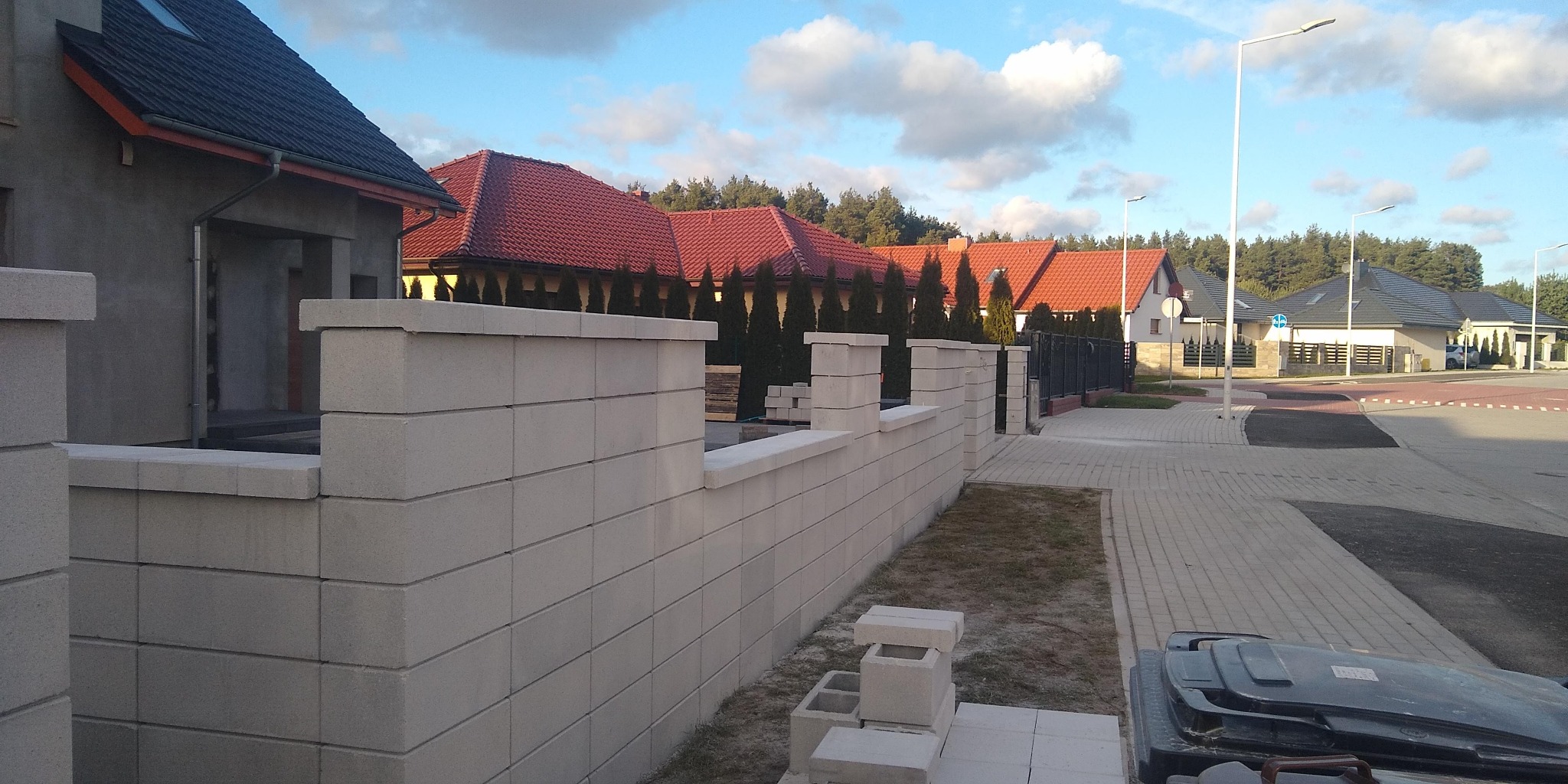 Nowoczesne osiedle z ogrodzeniem z betonowych bloczków, widoczne elementy w trakcie montażu, w tle domy z czerwonymi dachami i czarnym metalowym ogrodzeniem.