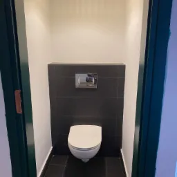 Wąska toaleta z czarnymi płytkami na podłodze i częściowo na ścianie, biała miska WC, chromowany przycisk spłuczki i żółta lampa wisząca z drewnianych listewek.