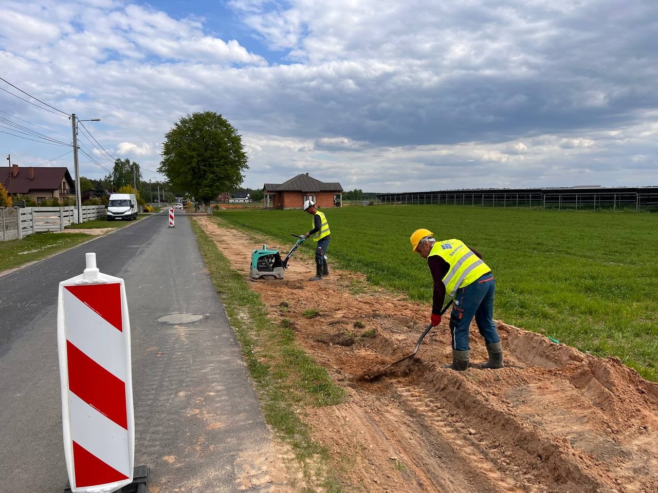 Dwaj pracownicy w kamizelkach odblaskowych wyrównują teren przy drodze za pomocą ubijarki i łopaty, w tle widoczne panele słoneczne na polu.