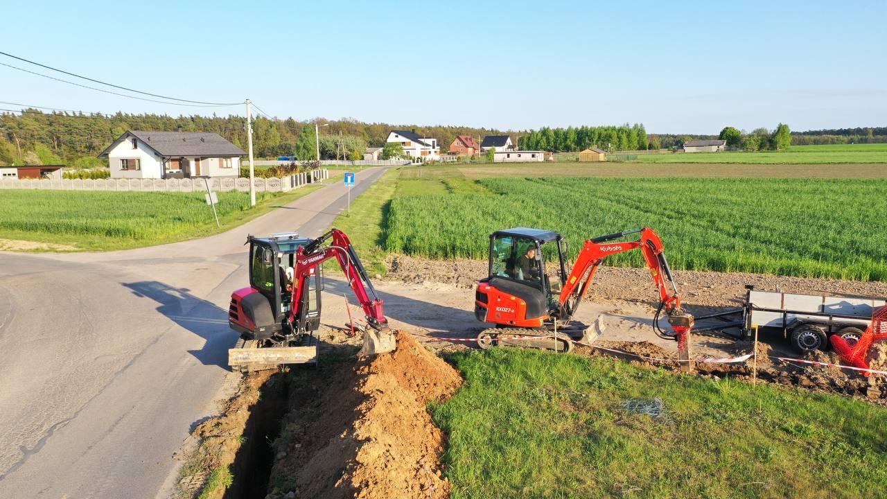 Dwa minikoparki Kubota i Yanmar pracują przy wykopie wzdłuż asfaltowej drogi, z widokiem na wiejski krajobraz z domami i polami w tle.