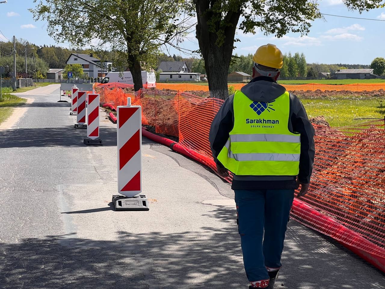 Pracownik w żółtej kamizelce z logo firmy 'Sarakhman Solar Energy' i żółtym kasku przechadza się po nowo wyasfaltowanej drodze, zabezpieczonej pomarańczową siatką i znakami ostrzegawczymi, w tle...