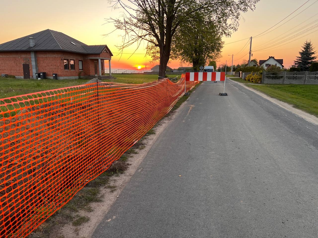 Pomarańczowa siatka zabezpieczająca wzdłuż drogi asfaltowej, w tle budynek w trakcie budowy i zachodzące słońce.