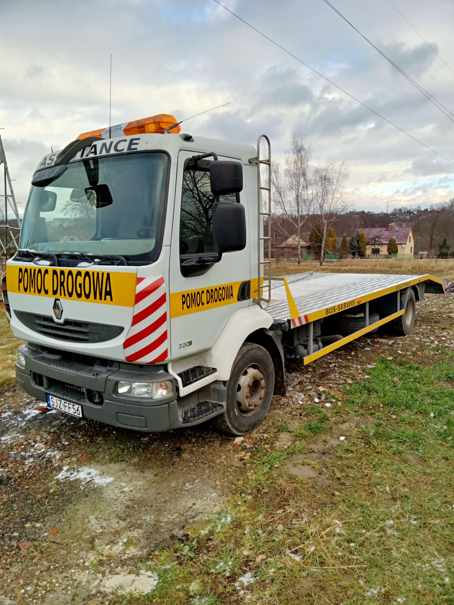 Biała laweta Renault z napisem 'Pomoc Drogowa' i żółtymi elementami stoi na błotnistym terenie z resztkami śniegu, widoczne budynki w tle.