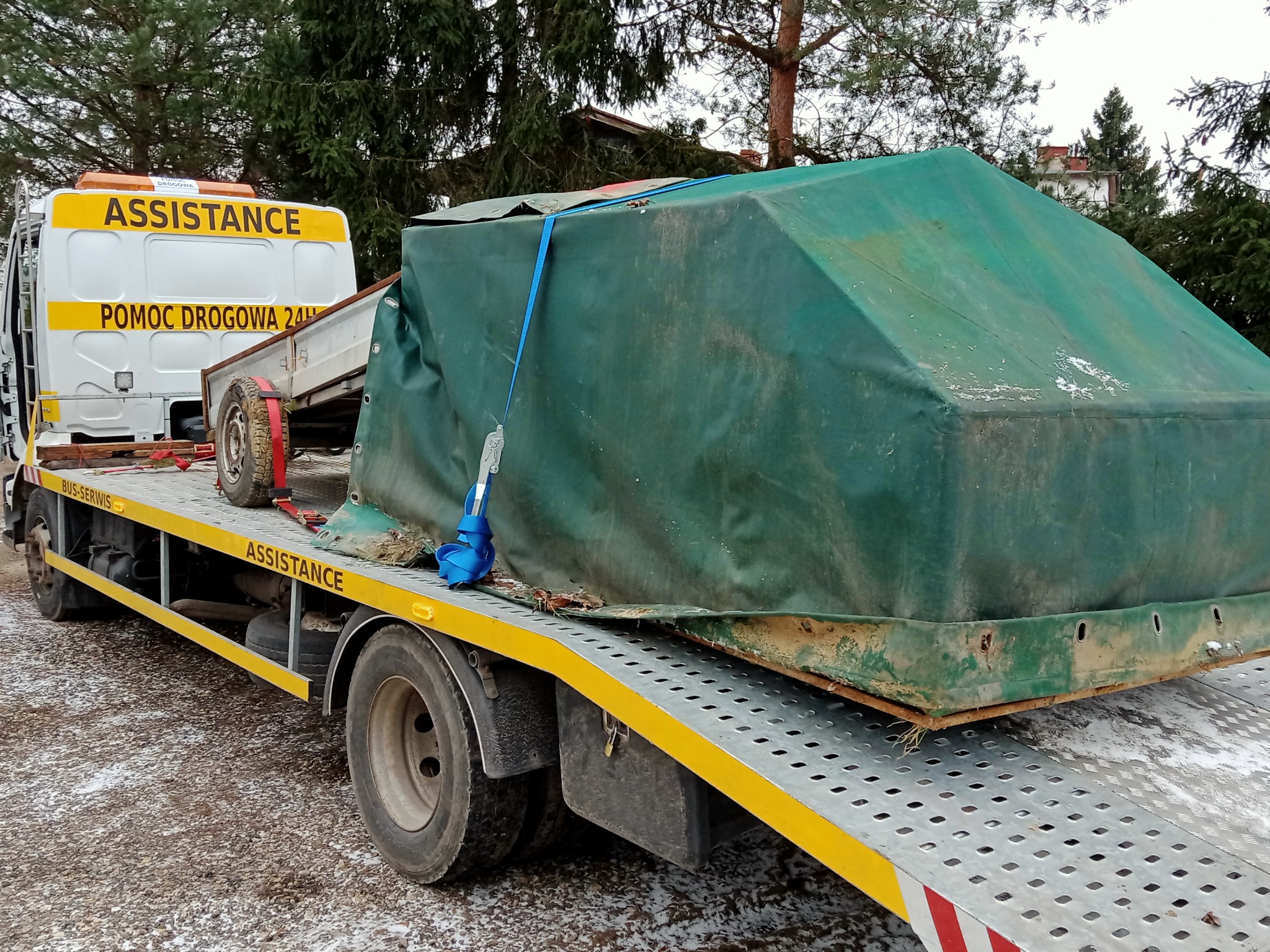 Laweta z napisem 'ASSISTANCE' i 'POMOC DROGOWA 24H' przewozi przyczepę okrytą zieloną plandeką, zabezpieczoną niebieskimi pasami.