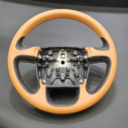 Odnowiona kierownica samochodowa z jasnobrązową skórą i czarnymi wstawkami, widoczne śruby montażowe i pomarańczowe szwy, na ciemnoszarym tle.