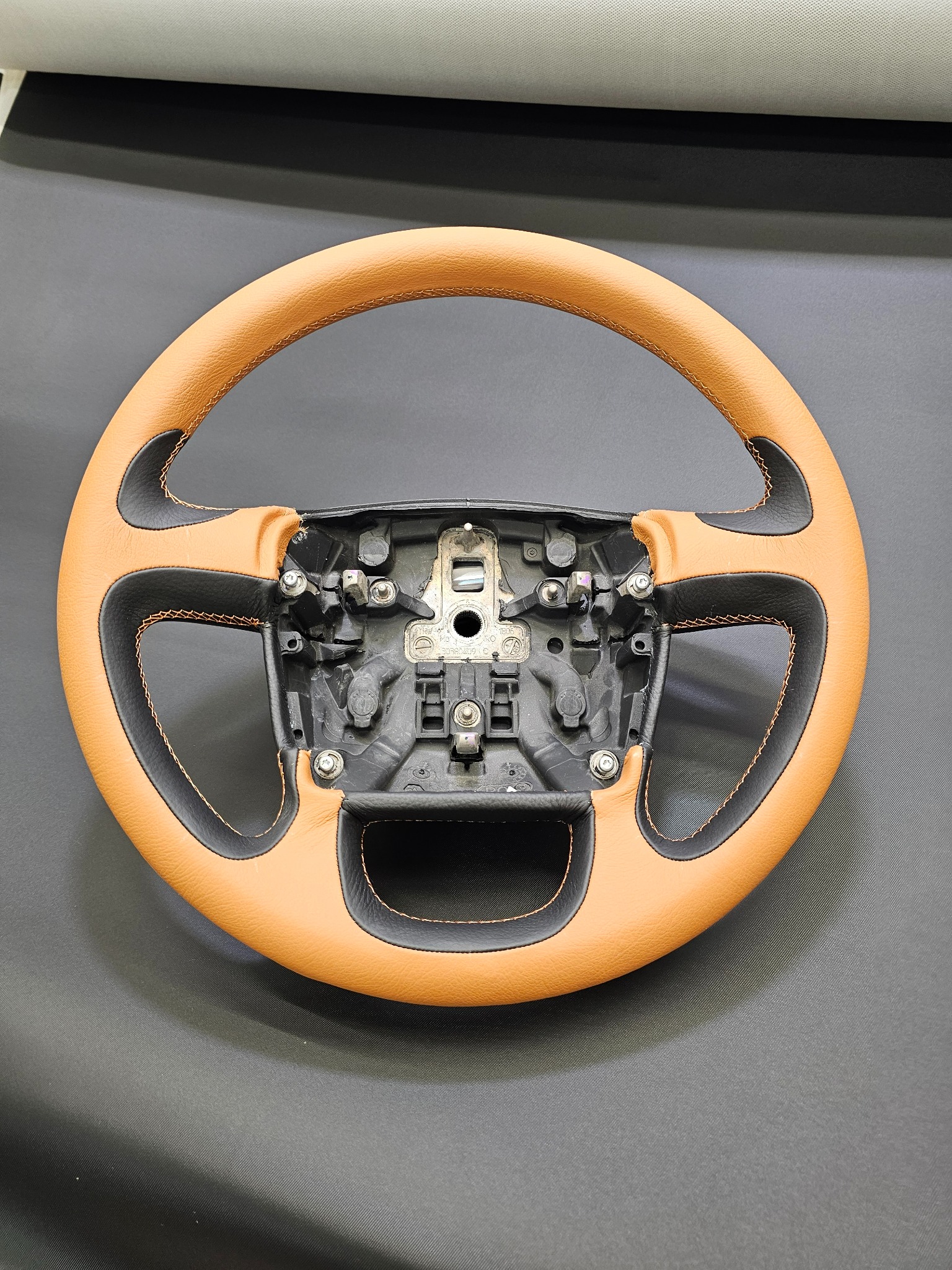 Odnowiona kierownica samochodowa z jasnobrązową skórą i czarnymi wstawkami, widoczne śruby montażowe i pomarańczowe szwy, na ciemnoszarym tle.