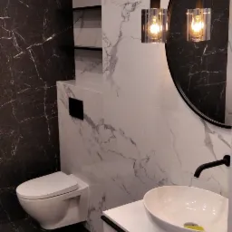 Nowoczesna łazienka z marmurowymi płytkami, wiszącą toaletą, okrągłym lustrem i designerskimi lampami wiszącymi z żarówkami Edisona.