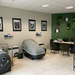 Wnętrze gabinetu z dwoma komorami do terapii hiperbarycznej, biurkiem, krzesłami i dekoracjami roślinnymi na ścianie w kolorze butelkowej zieleni.