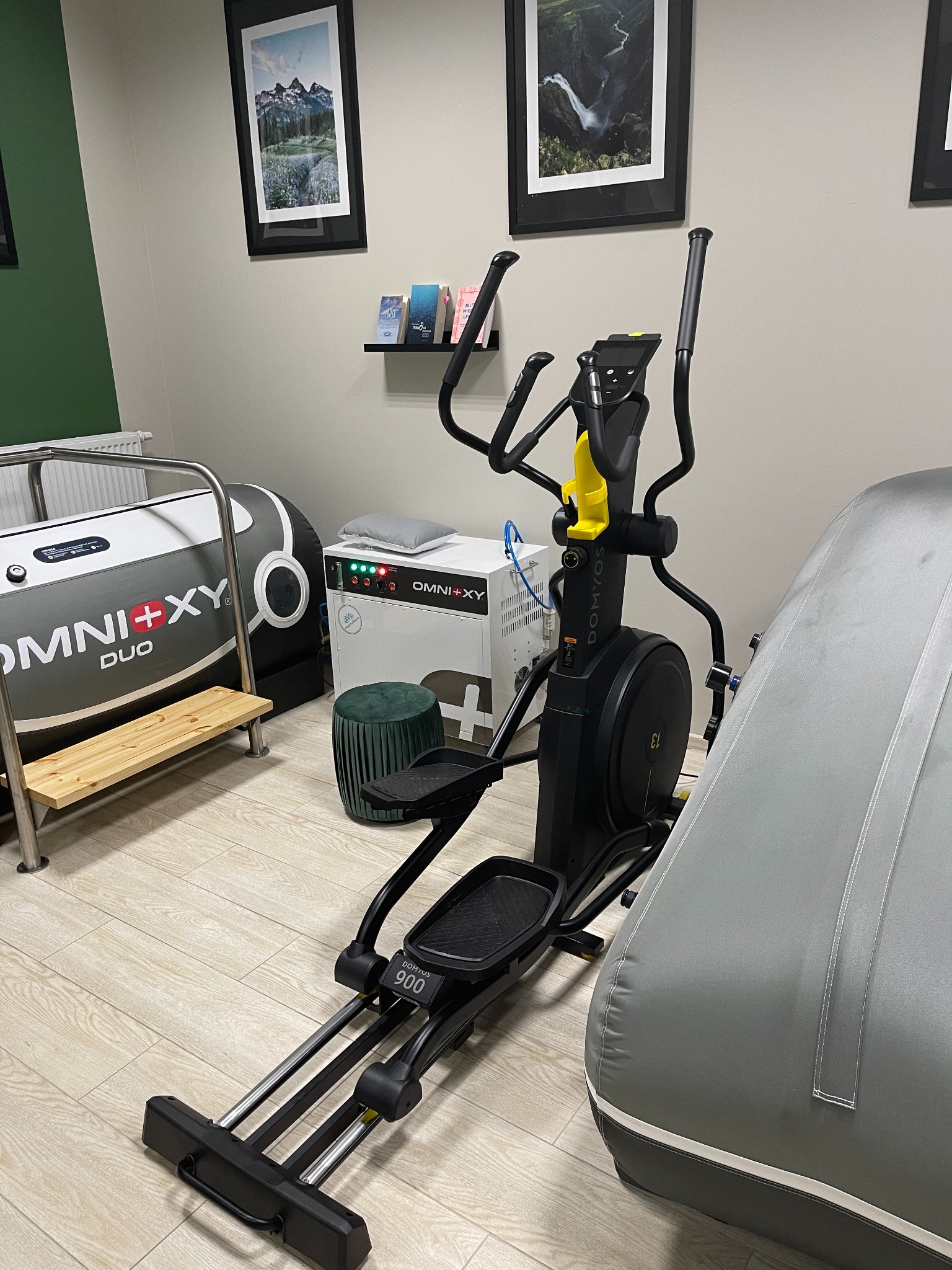 Wnętrze gabinetu rehabilitacyjnego z czarnym orbitrekiem Domynos 900, urządzeniem do terapii Omni+XY Duo oraz obrazami gór i wodospadu w czarnych ramkach na ścianie.