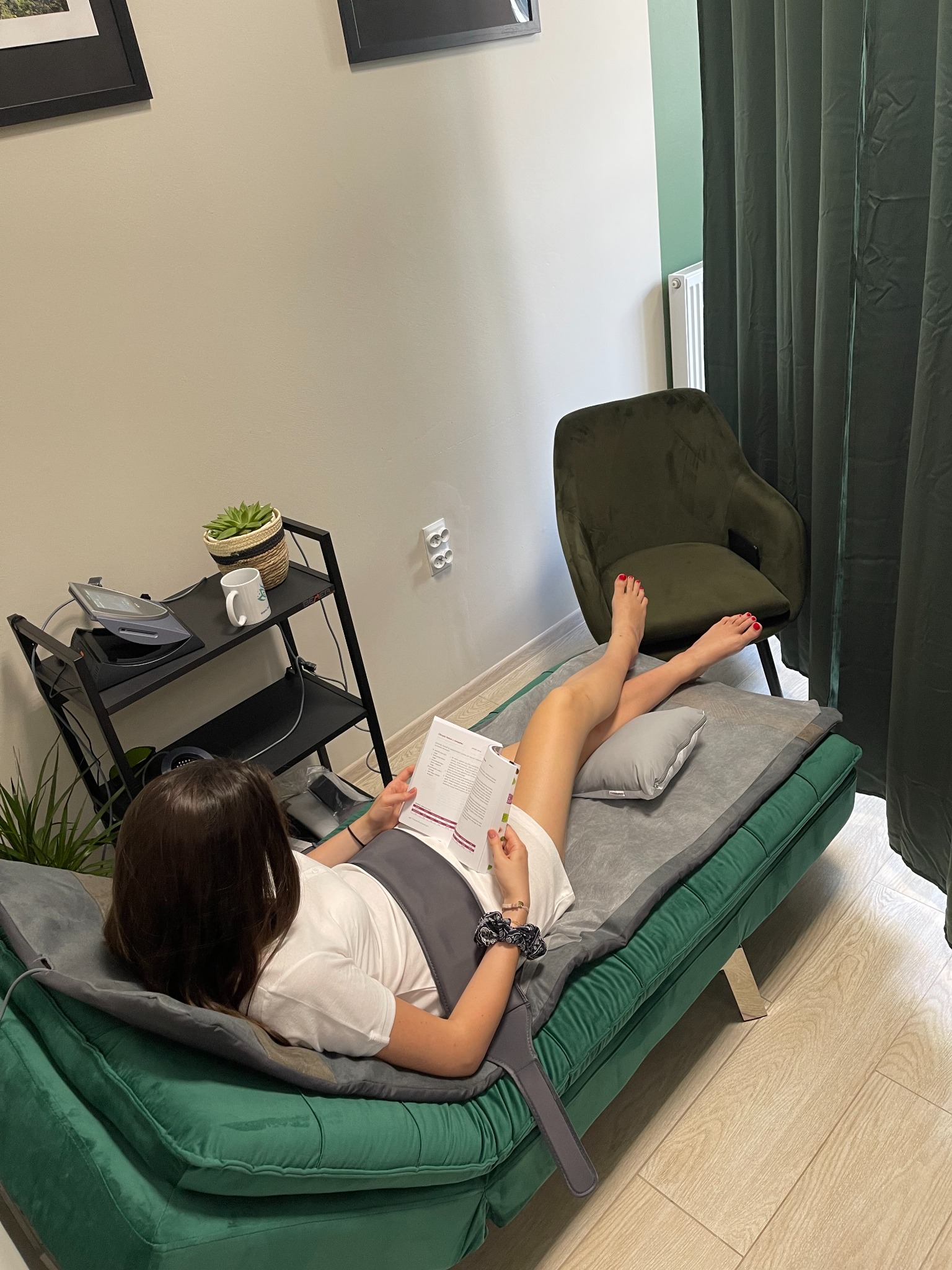 Kobieta leży na zielonym tapczanie z szarym materacem, czytając dokument, w tle fotel, zasłony i półka z urządzeniem do terapii.