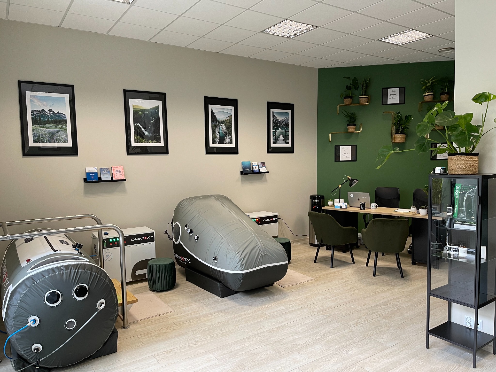 Wnętrze gabinetu z dwoma komorami do terapii hiperbarycznej, biurkiem, krzesłami i dekoracjami roślinnymi na ścianie w kolorze butelkowej zieleni.