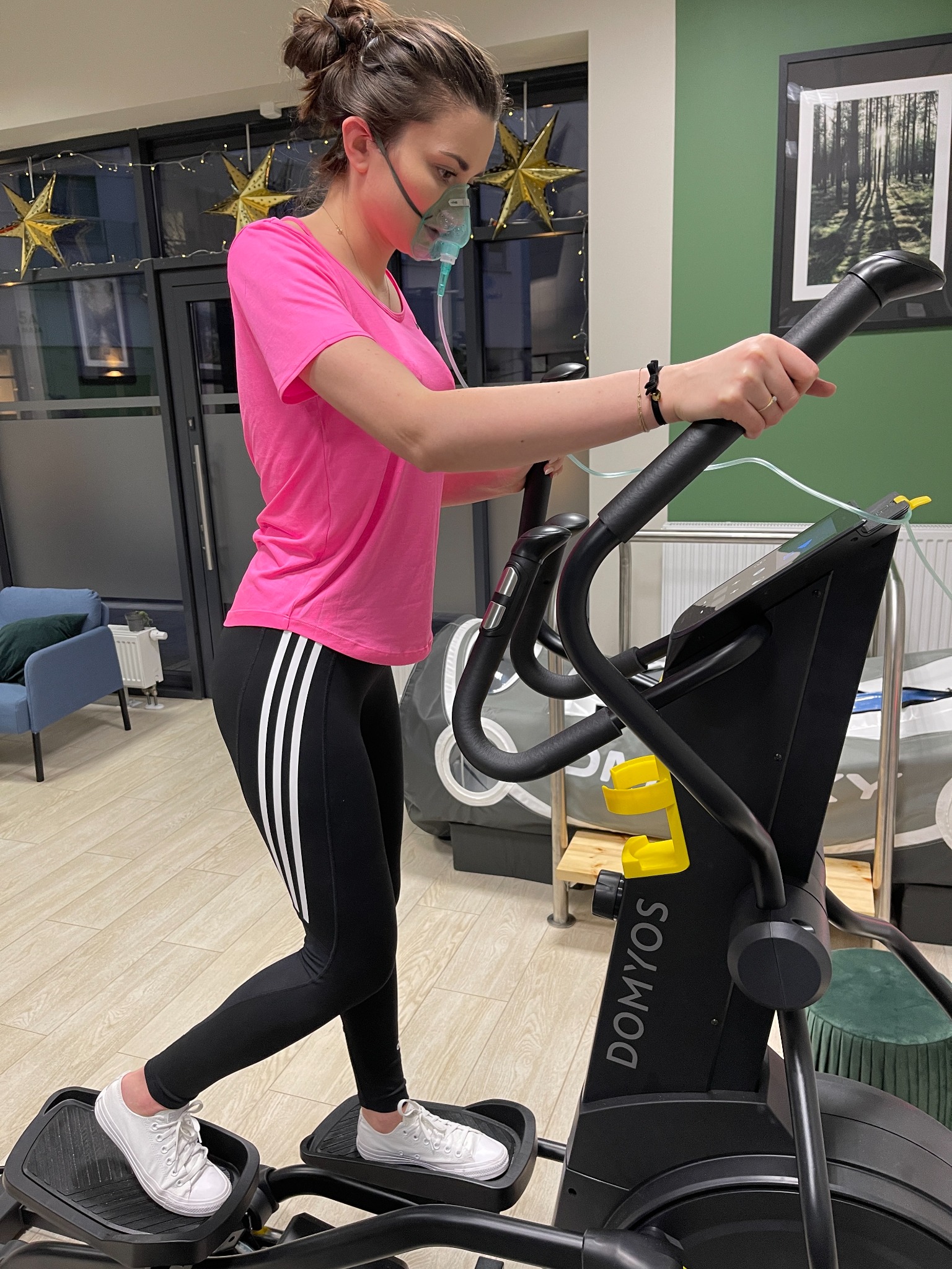 Kobieta w różowej koszulce i czarnych legginsach ćwiczy na orbitreku marki Domyos, ma założoną maskę tlenową, w tle widoczne dekoracje świąteczne i obraz na ścianie.