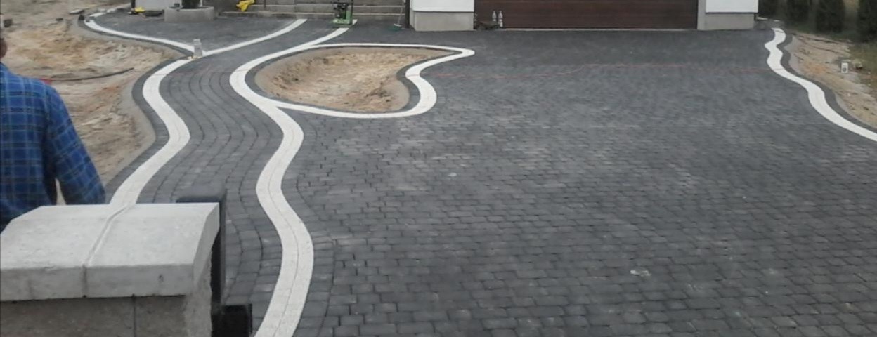 Ukończony podjazd z ciemnej kostki brukowej z falującymi pasami jasnej kostki, otaczający rabatę ziemną i prowadzący do garażu.