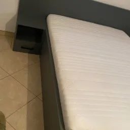 Szare łóżko z wbudowaną szafką nocną i białym materacem, ustawione na podłodze z płytek ceramicznych.