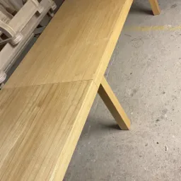 Długi blat drewniany z widocznym łączeniem, na ukośnych nogach, w warsztacie stolarskim, z elementami krzeseł w tle.