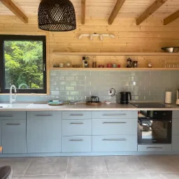 Jasna kuchnia w drewnianym domu z niebieskimi frontami szafek, szarą glazurą i drewnianym sufitem z belkami.