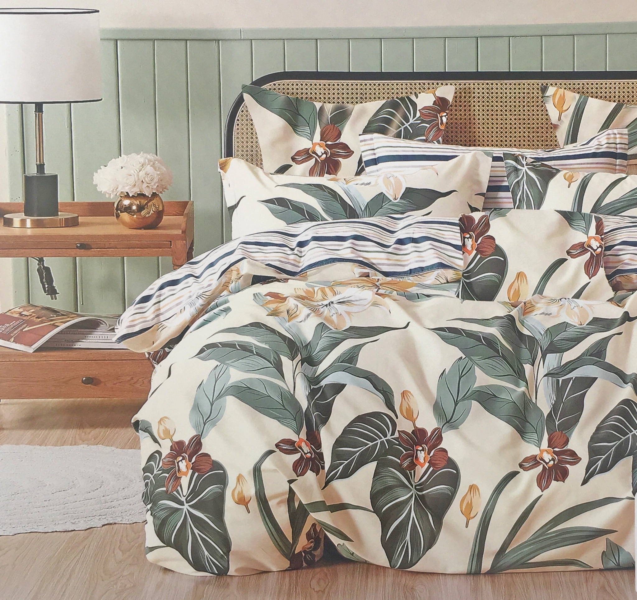Komplet pościeli z motywem tropikalnych liści i kwiatów na łóżku z rattanowym zagłówkiem, obok drewniany stolik nocny z lampką i wazonem z białymi kwiatami.