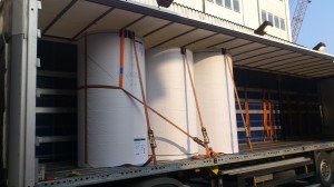 Wnętrze naczepy ciężarówki z trzema dużymi rolami białego papieru zabezpieczonymi pomarańczowymi pasami transportowymi. Widoczna metalowa konstrukcja naczepy oraz oświetlenie. W tle fragment...