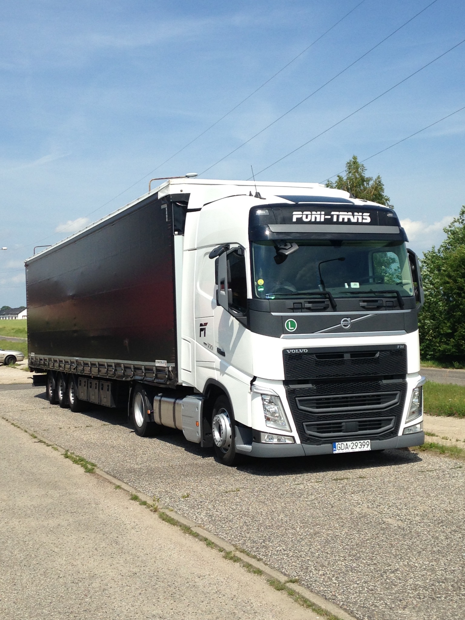 Biała ciężarówka Volvo z czarną naczepą, z napisem 'PONI-TRANS' nad przednią szybą, zaparkowana na placu w słoneczny dzień.