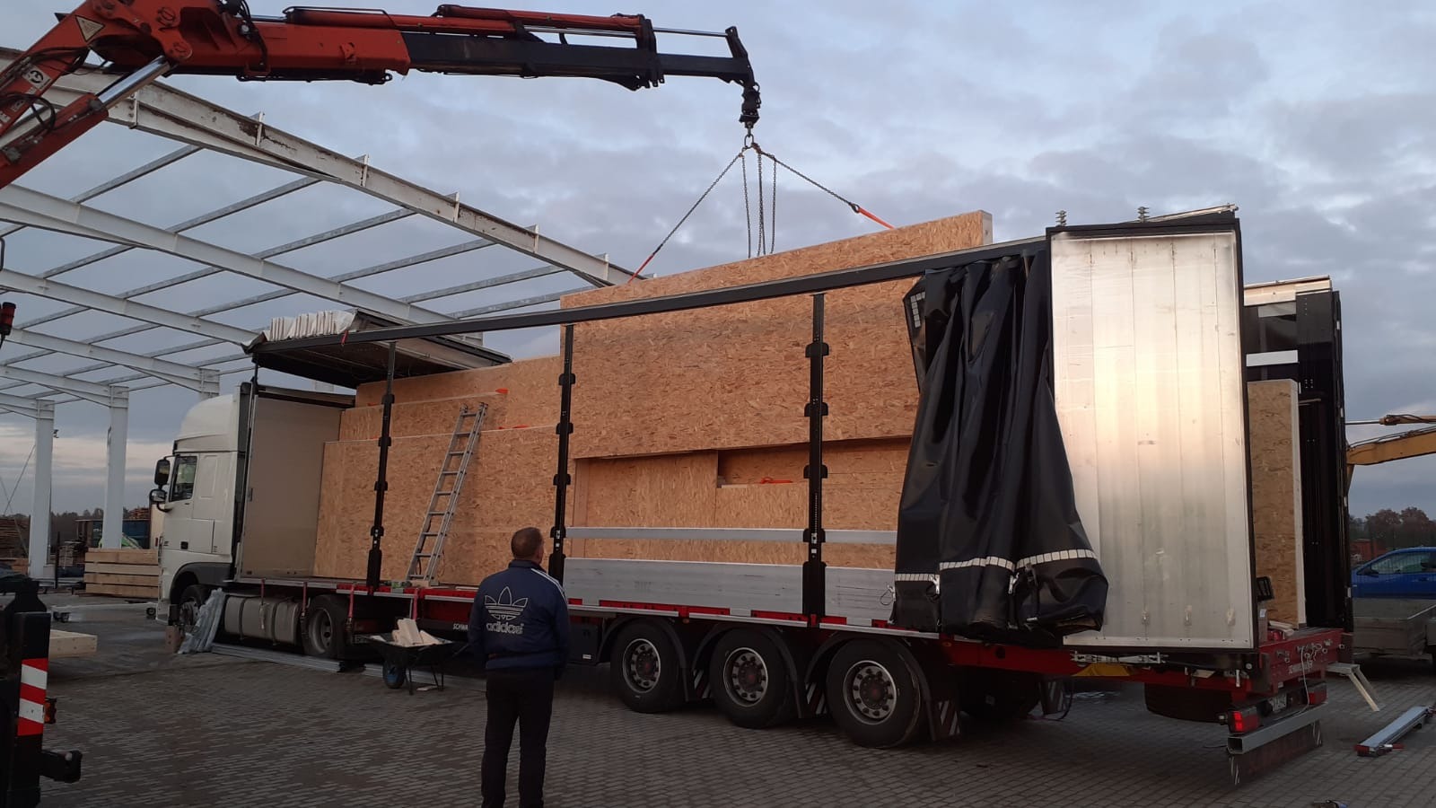 Ciężarówka z otwartą naczepą załadowana płytami OSB, rozładunek przy pomocy dźwigu, widoczna drabina oparta o ładunek.