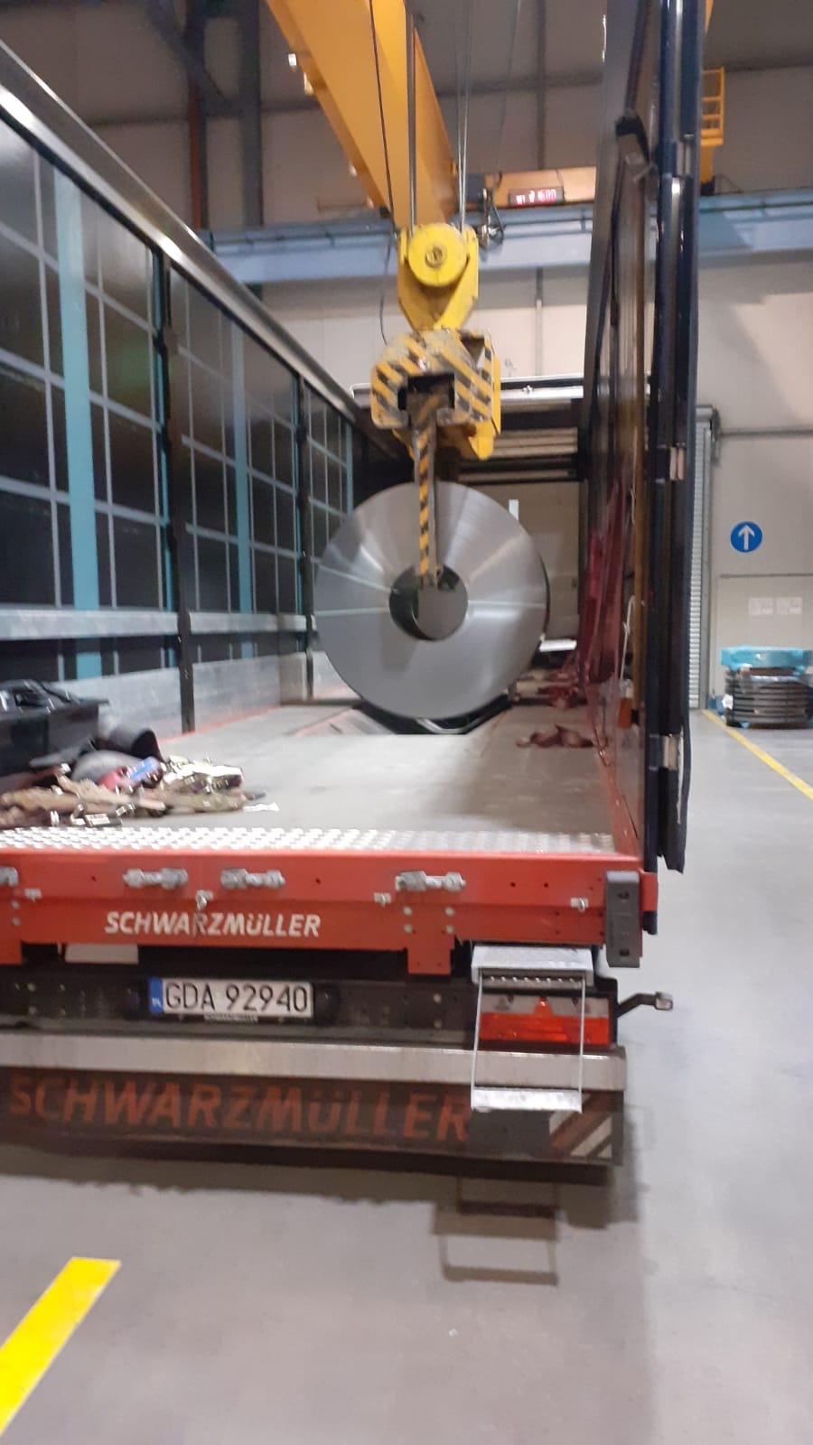Załadunek stalowej rolki blachy do naczepy ciężarówki za pomocą suwnicy w hali przemysłowej. Widoczne logo Schwarzmüller na burcie.