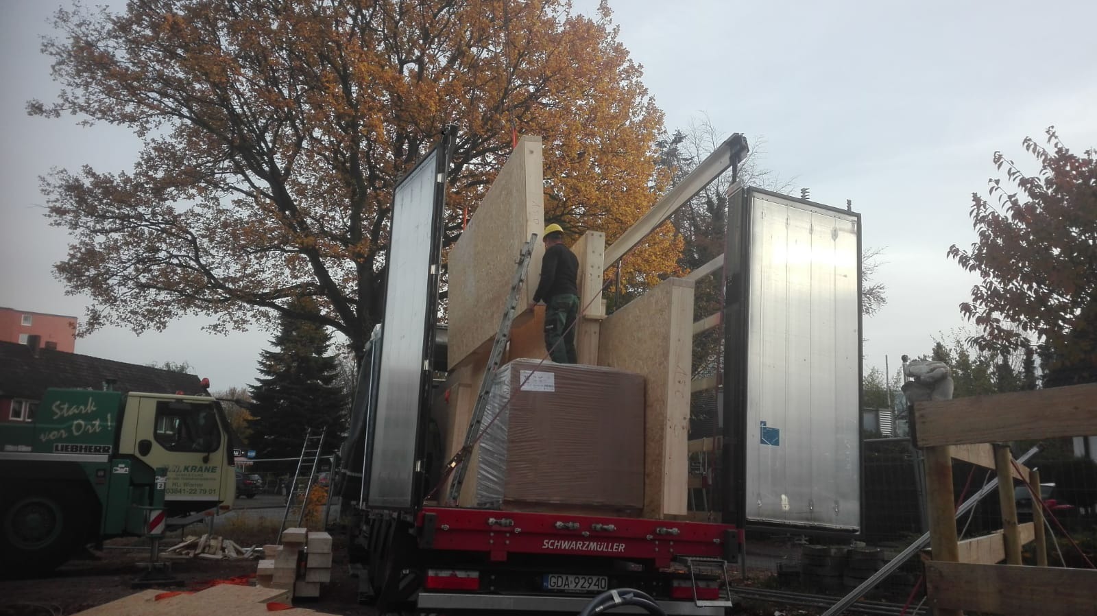 Pracownik w kasku na drabinie wewnątrz naczepy ciężarówki, załadunek prefabrykowanych elementów konstrukcyjnych z drewna, na zewnątrz widoczny dźwig budowlany.